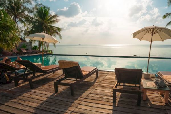 A tranquil poolside scene with sun loungers, umbrellas, and a view of the ocean, surrounded by palm trees under a partly cloudy sky.