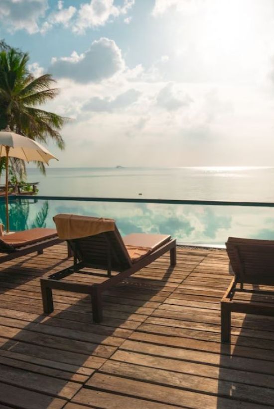 A serene poolside scene with lounge chairs, umbrellas, palm trees, and an ocean view under a partly cloudy sky at sunset.