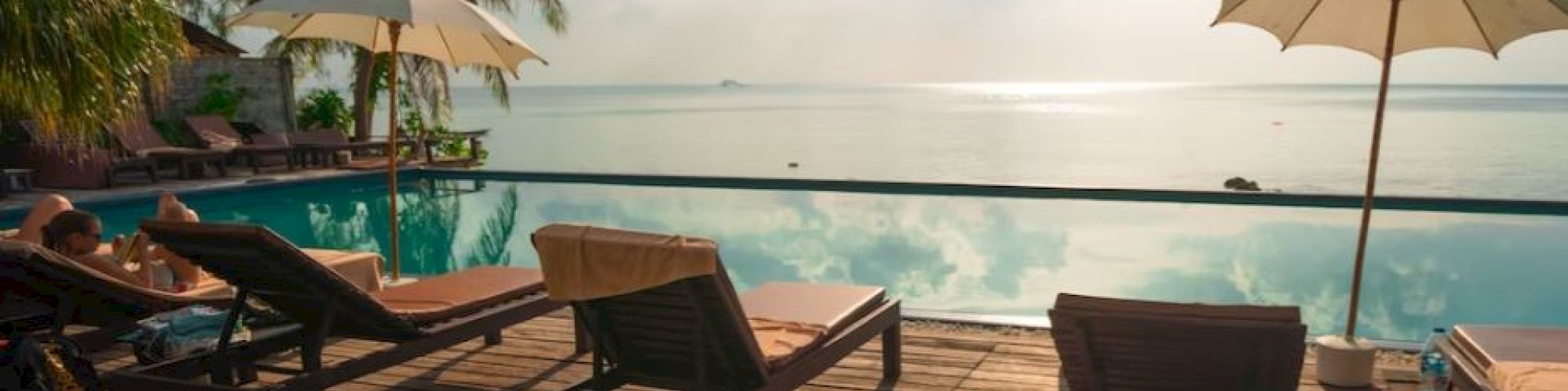 Lounge chairs and umbrellas by a serene pool with a picturesque ocean view and palm trees under a bright sky.