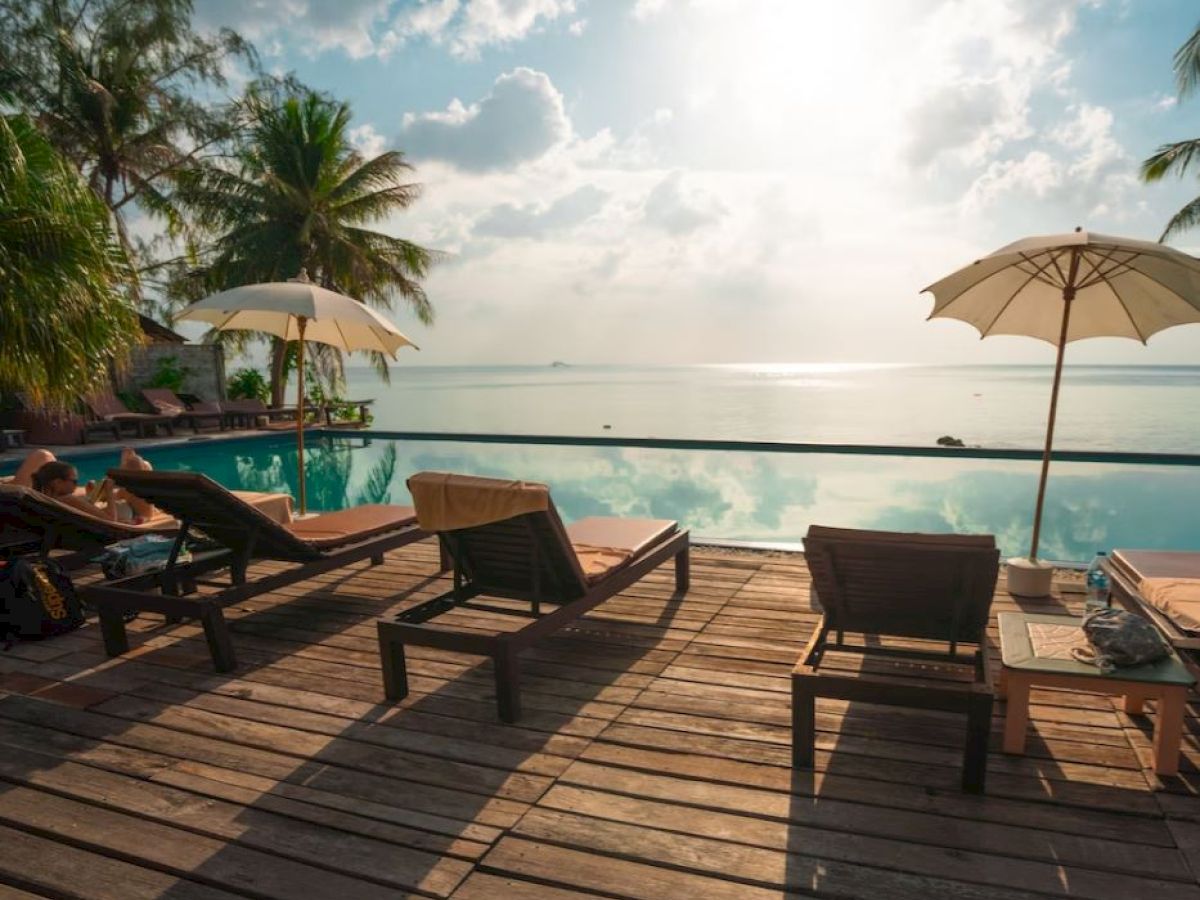 A serene poolside scene with lounge chairs, umbrellas, and a view of the ocean horizon under a sunny, partly cloudy sky.