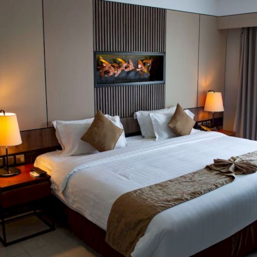 The image shows a cozy hotel room with a large bed, two bedside lamps, and decorative pillows, creating a warm and inviting atmosphere.