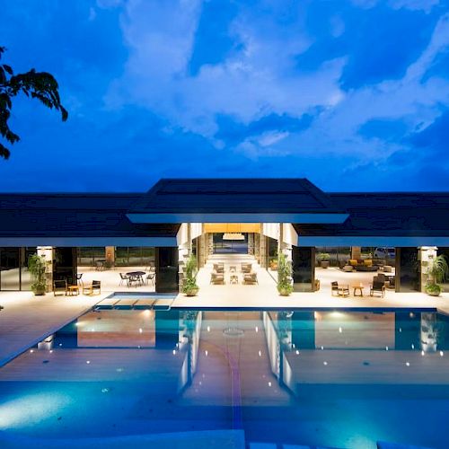 A modern building with an illuminated swimming pool at dusk, surrounded by seating areas under a clear, deep blue sky.