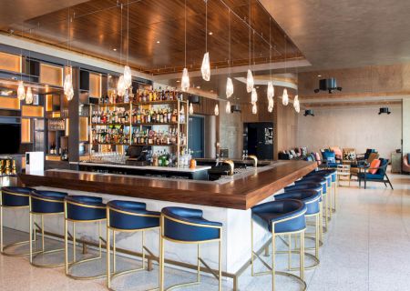 The image shows a modern bar with a central counter, stylish chairs, pendant lights, and a wide selection of bottles on display.