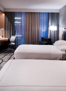 The image shows a modern hotel room with two beds, a TV, desk, chair, and stylish lighting. The decor is sleek with patterned walls and curtains.