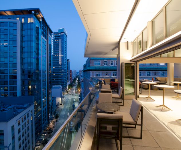 A modern urban balcony with seating overlooks a city street lined with tall buildings at twilight, featuring large windows and city lights.