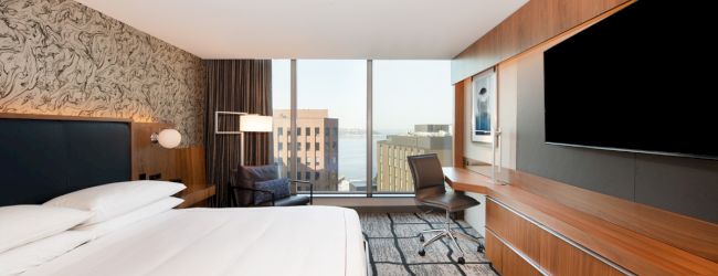 A modern hotel room features a bed, desk, chair, large TV, and a window with a city view in the distance.