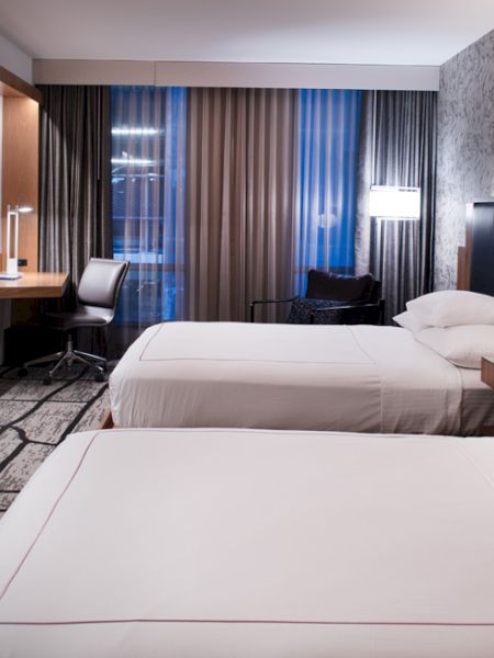 The image shows a modern hotel room with two beds, a TV, a desk, and contemporary lighting in a stylish setting.