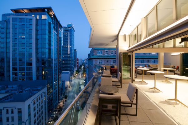 A modern rooftop patio with tables and chairs overlooking a cityscape at dusk, featuring glass buildings and a street view below.