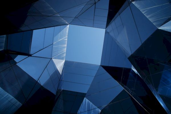 The image shows modern architectural structures with reflective glass panels, forming a geometric pattern against a blue sky.