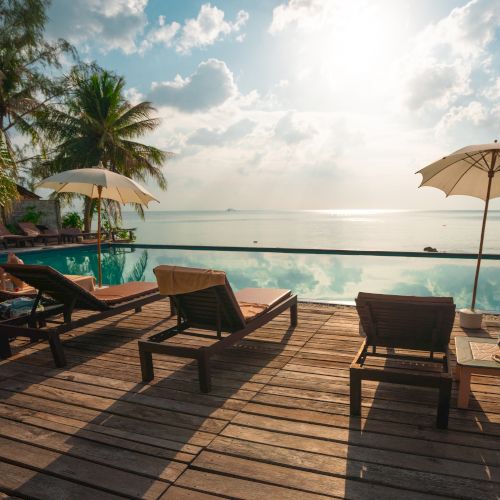 Sun loungers on a wooden deck overlook an infinity pool and tranquil ocean, with palm trees and umbrellas under a bright, partly cloudy sky.