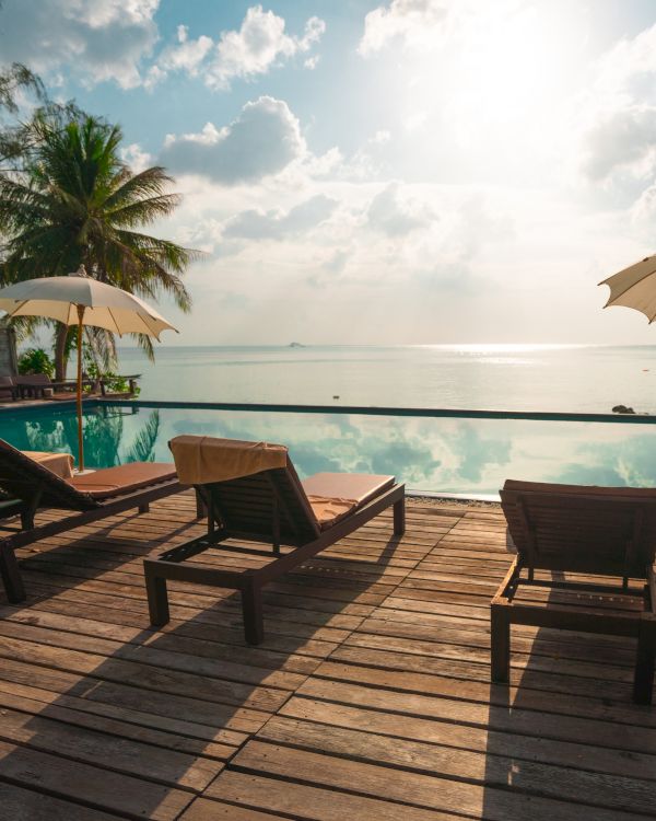 Sun loungers on a wooden deck overlook an infinity pool and tranquil ocean, with palm trees and umbrellas under a bright, partly cloudy sky.