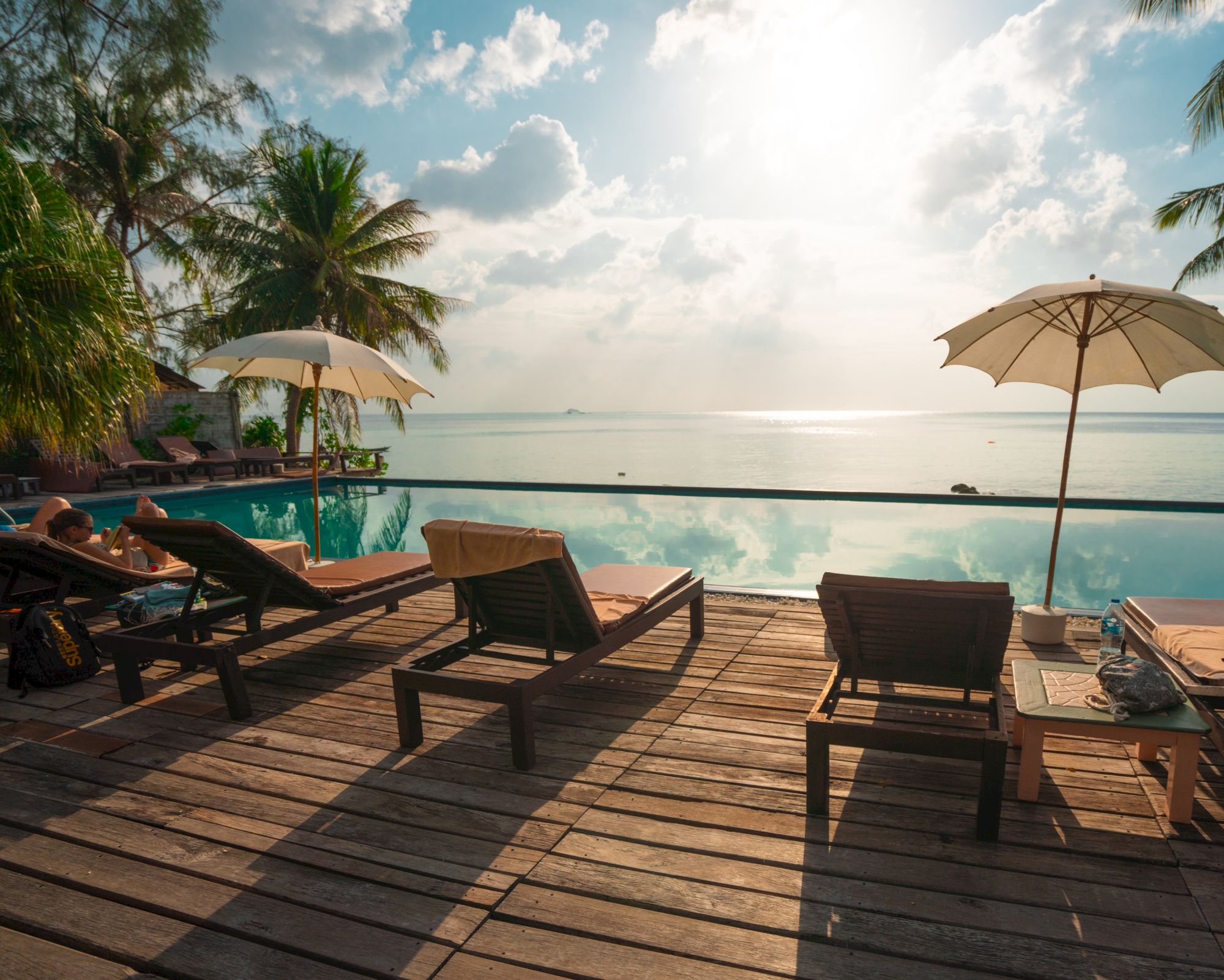 A serene beach scene with lounge chairs, umbrellas, and palm trees facing a calm ocean under a partly cloudy sky at sunrise or sunset.