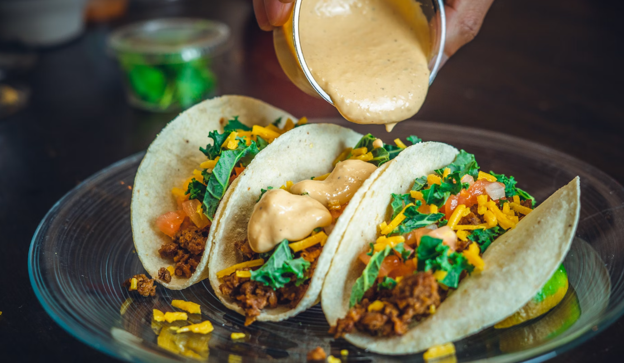 Three tacos with meat, shredded cheese, kale, and tomatoes are being topped with a creamy sauce on a glass plate.