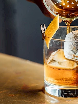 A hand pours a brown liquid into a glass with ice and a lemon twist on a wooden surface.