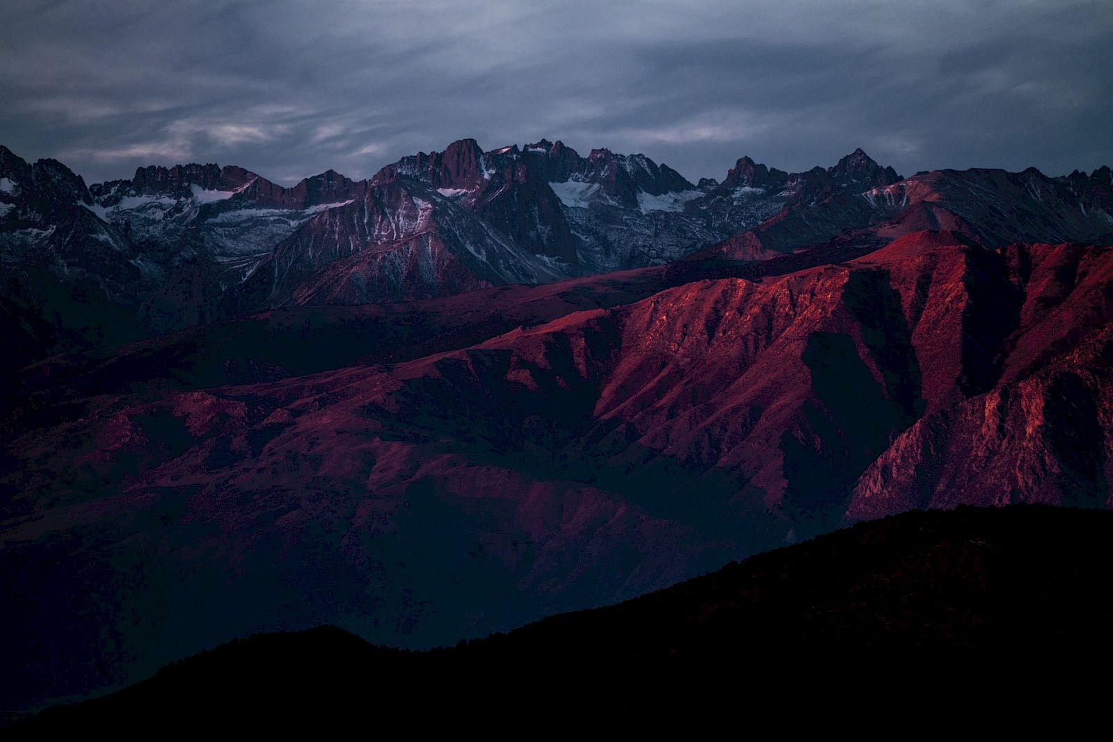 A mountain range bathed in a reddish hue under a cloudy sky during sunset or sunrise, creating a dramatic landscape scene.