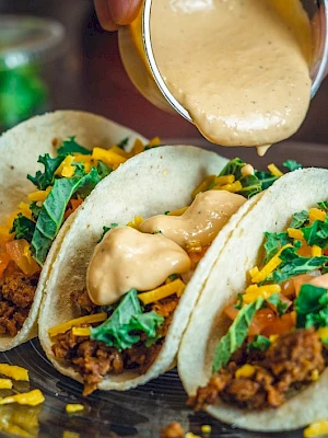 Three tacos with ground beef, cheese, tomatoes, and greens are being topped with creamy sauce on a plate.