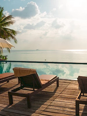 Poolside scene with lounge chairs, umbrellas, palm trees, and an ocean view under a cloudy sky.