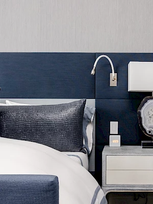A modern bedroom features a bed with blue and white bedding, a side table with a lamp, and a wall-mounted reading light.
