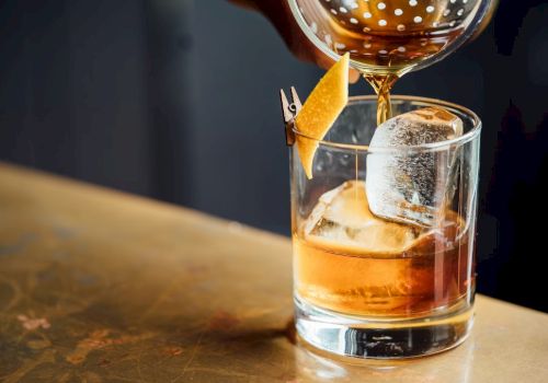A cocktail is being poured into a glass with ice and a citrus peel garnish on a metal bar counter.