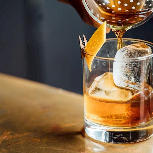 A cocktail is being poured into a glass with ice and a citrus peel garnish on a metal bar counter.