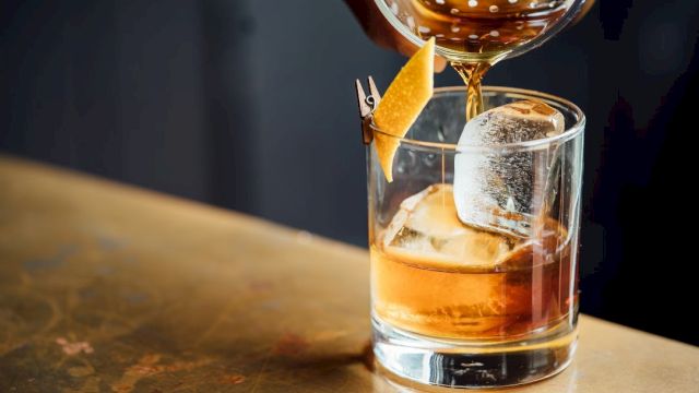 A cocktail is being poured into a glass with ice and a citrus peel garnish on a metal bar counter.