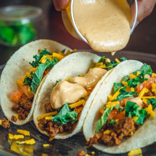 Three tacos with ground beef, cheese, lettuce, and tomatoes are being topped with a creamy sauce on a plate.