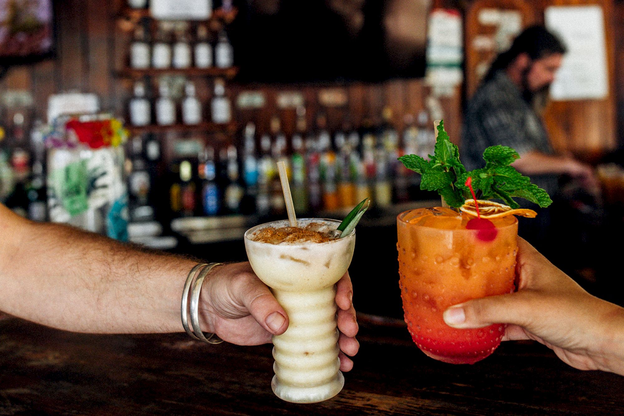 Two people hold colorful cocktails at a bar; one drink is creamy with a straw, the other is orange with mint and garnishes.