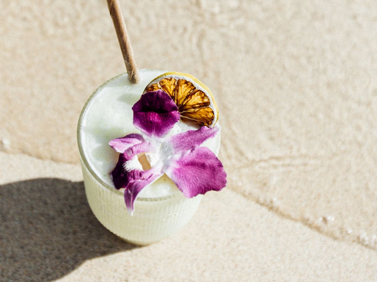 A tropical drink garnished with a purple flower and a slice of dried citrus, placed on a sandy surface with a wooden straw.