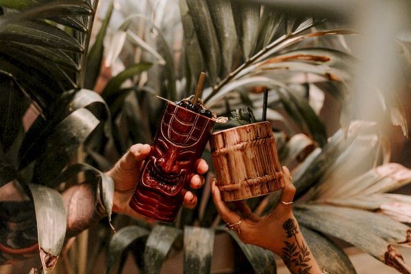 Two hands holding tiki-style mugs with straws, surrounded by tropical foliage, creating a festive and exotic vibe.