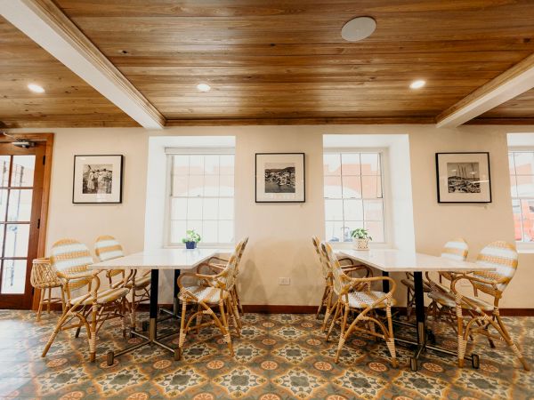 The image shows a room with rattan chairs around white tables, patterned tile flooring, and framed photos on the wall beneath a wooden ceiling.