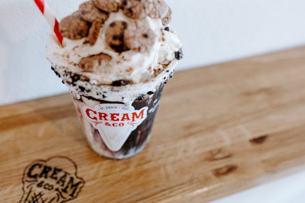 A milkshake topped with whipped cream and cereal is in a cup labeled "Cream & Co," on a wooden surface with the same logo.
