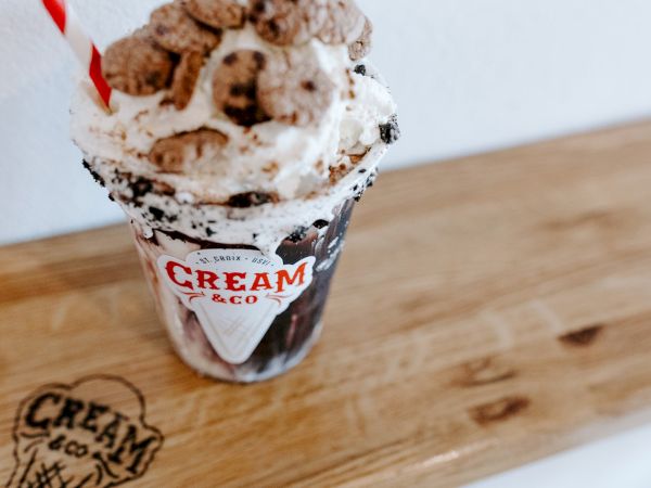 A milkshake topped with whipped cream and cereal is in a cup labeled "Cream & Co," on a wooden surface with the same logo.