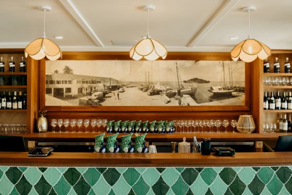 A stylish bar with hanging lights, a vintage harbor photo, wine bottles, glasses, and green-patterned tiles on the front counter.