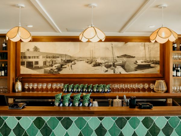 A stylish bar with hanging lights, a vintage harbor photo, wine bottles, glasses, and green-patterned tiles on the front counter.
