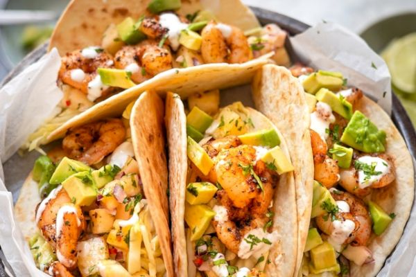 The image shows three delicious shrimp tacos with avocado, cheese, and sauce. They are garnished with cilantro in a bowl.