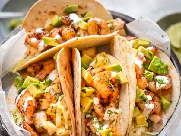 The image shows three delicious shrimp tacos with avocado, cheese, and sauce. They are garnished with cilantro in a bowl.