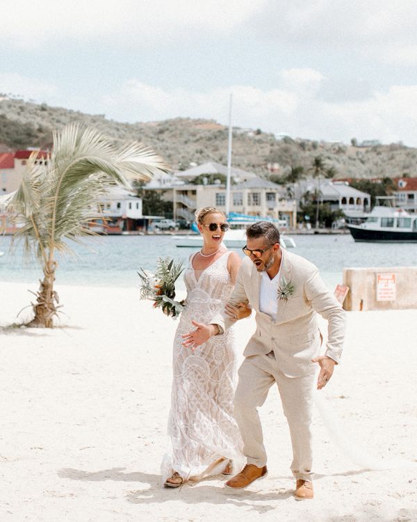 A couple in formal attire is playfully posing on a beach with boats and buildings in the background, under a partly cloudy sky.