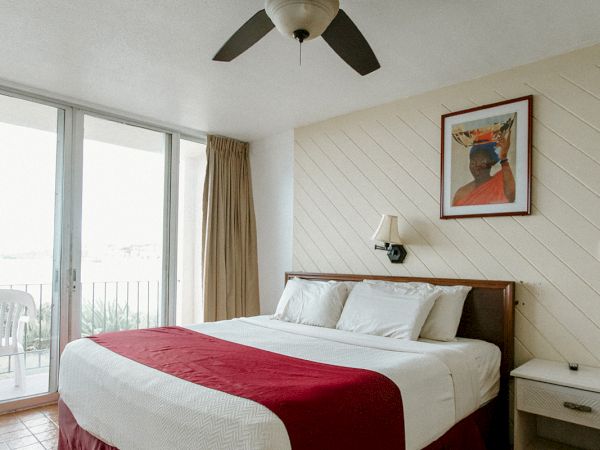 A neatly arranged hotel room with a large bed, red bedspread, side tables, and a ceiling fan, with a painting and window in the background.