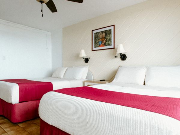 The image shows a hotel room with two beds, red bedspreads, a ceiling fan, and a framed artwork on the wall above the beds.