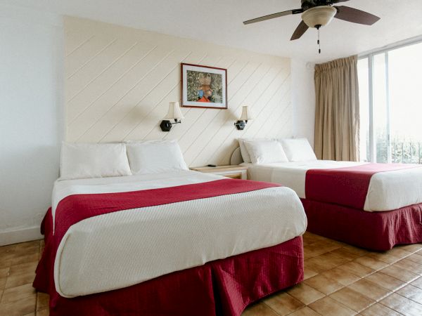 A hotel room with two beds, red bed skirts, wall lamps, a ceiling fan, and a window with curtains. A picture hangs above the beds.
