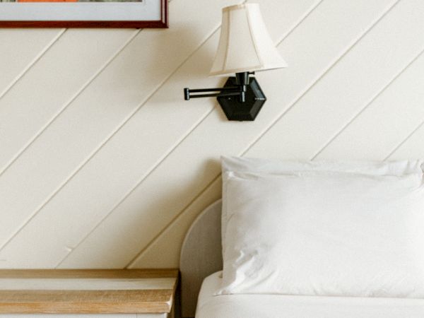 A cozy bedroom scene with a bed, a wooden nightstand, a wall-mounted lamp, and a framed artwork above the nightstand.