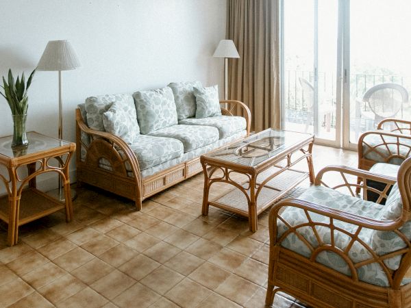A living room with wicker furniture, cushioned seats, a glass-top table, lamps, and a plant, with tiled flooring and large windows.
