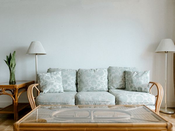 A living room with a sofa, glass coffee table, two lamps, and a plant on a side table against a plain wall.