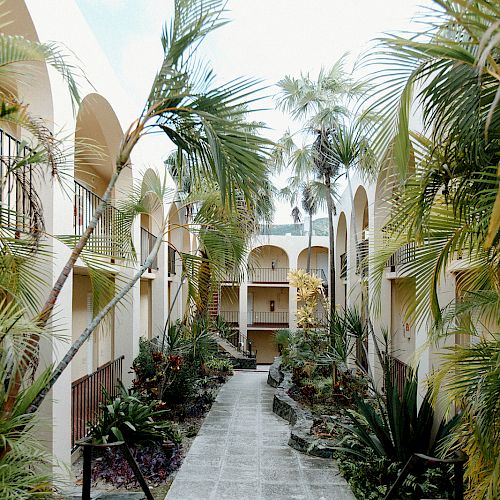 A courtyard with arched walkways, lush palm trees, and a pathway leading through the center, surrounded by a serene, tropical atmosphere.