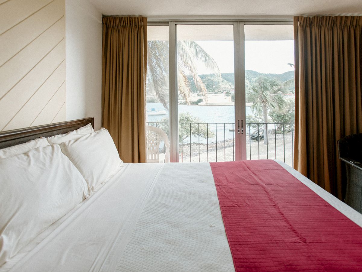 A bright bedroom with a large bed, red runner, and a window view of palm trees and water outside, covered by tan curtains.