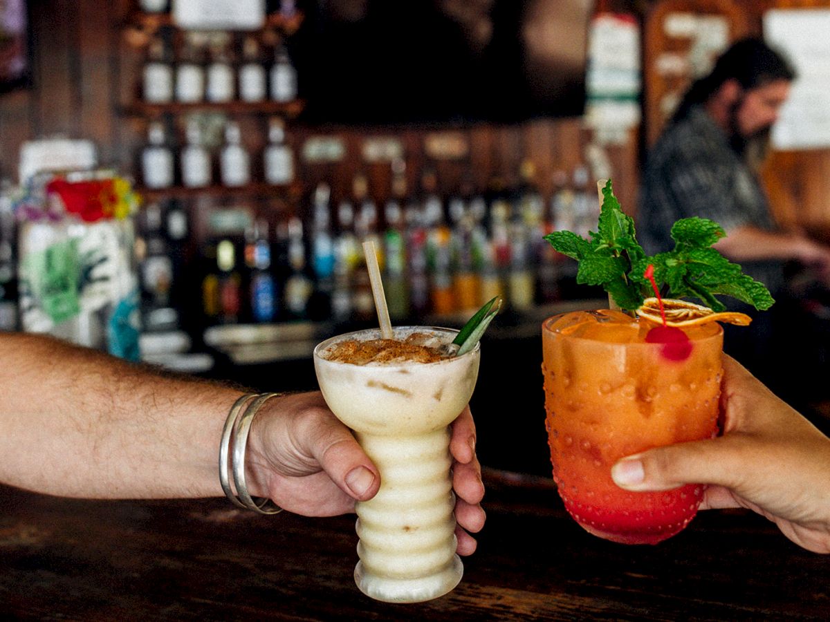 Two people are holding tropical cocktails at a bar, with blurred bottles in the background.
