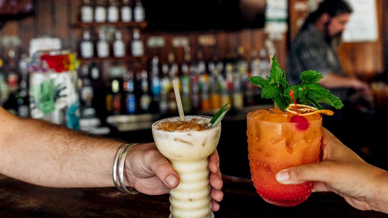 Two people are holding tropical cocktails at a bar, with blurred bottles in the background.