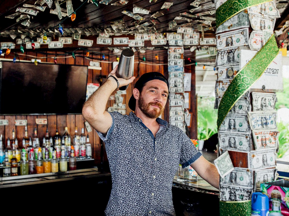 A person stands in a bar decorated with dollar bills on the ceiling and walls, holding a bill. Bottles and other items are visible behind.