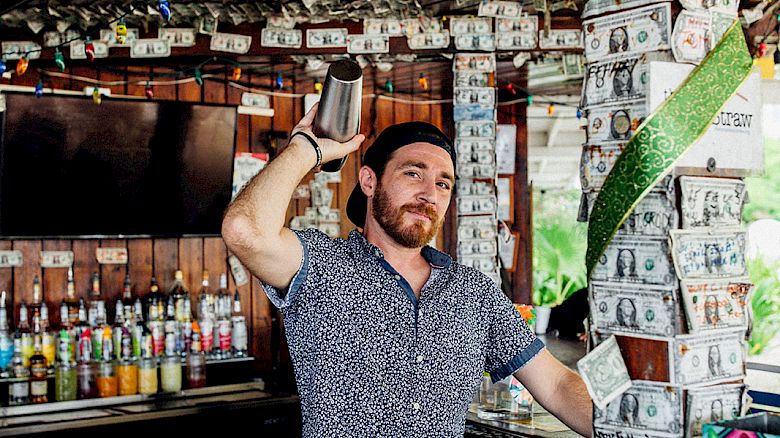 A person stands in a bar decorated with dollar bills on the ceiling and walls, holding a bill. Bottles and other items are visible behind.