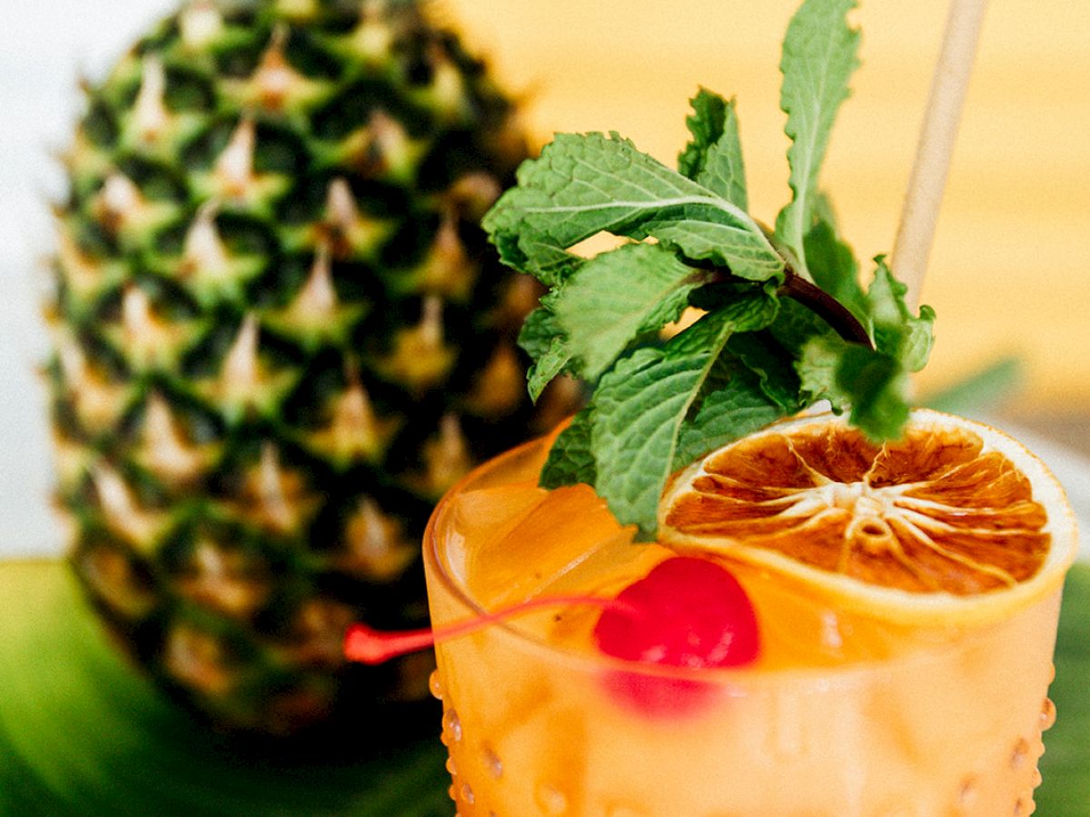 A tropical cocktail garnished with mint, orange slice, and cherry sits next to a pineapple on a banana leaf.